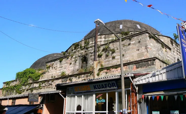 A dilapidated old stone building with a dome roof and overgrown vegetation stands as a testament to the rich history you’ll encounter when you visit Turkey. Below it, a modern street features shops, including a barber shop named "Kuaför." Colorful pennant banners are strung across the street under a clear blue sky.