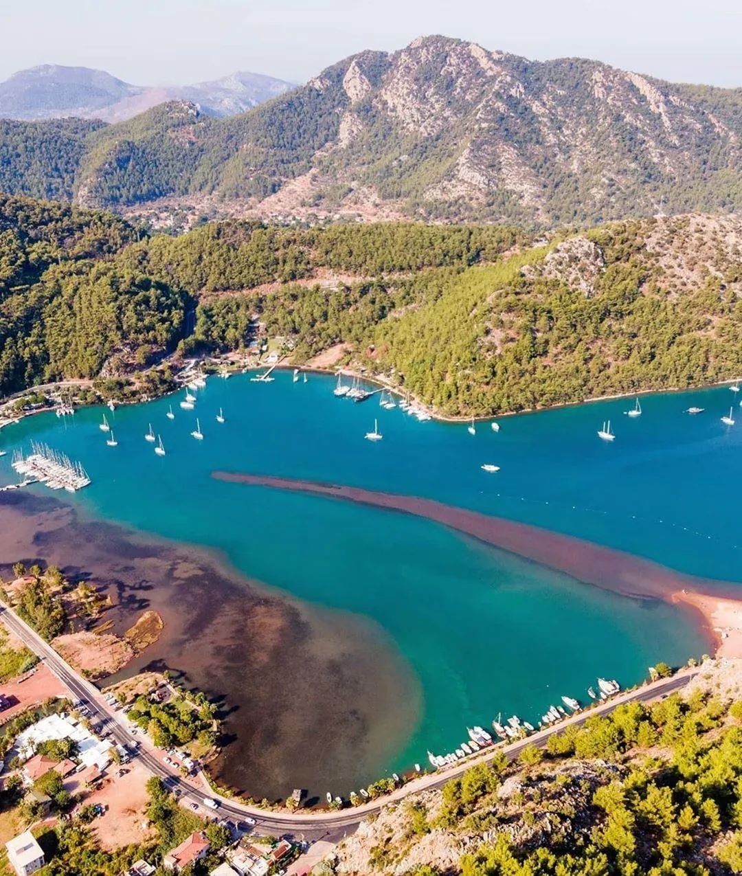 Scenic view of Kızkumu, Turkey, showcasing the unique 600-meter shallow water pathway where people are walking on water, with a statue of the girl from the legend in the foreground, lush green hills, and a clear blue sky in the background.